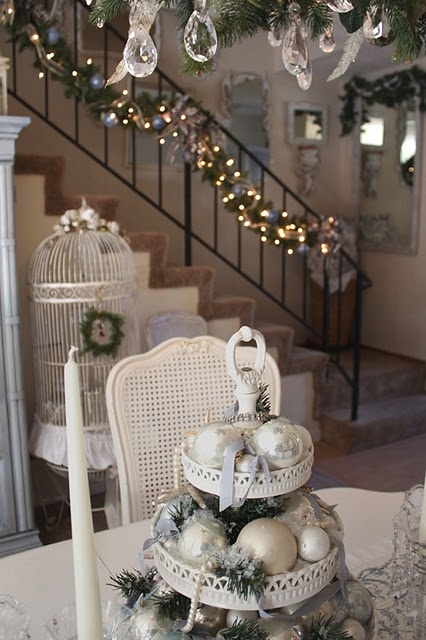 a white tiered stand with evergreens, white, pearly and silver Christmas ornaments, a white cage with a green wreath and a thin and tall candle