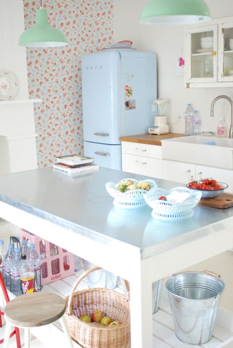 a cute vintage kitchen with floral wallpaper, white cabinets, a fireplace, a large table kitchen island, a blue fridge, mint green pendant lamps and touches of pink