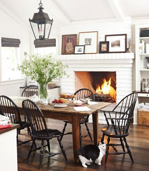 a bright white farmhouse dining space with a white brick fireplace, a wooden table and black chairs, artworks and lanterns