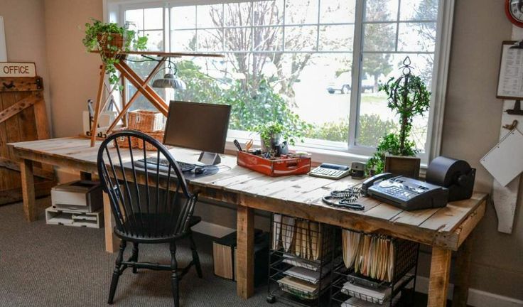 a farmhouse home office with a super long wooden desk with open storage units, a vintage chair, potted greenery and cool relaxing views