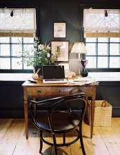 a vintage farmhouse home office with black walls, a wooden desk and a black chair, woven shades and a basket for trash
