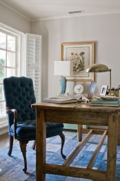 a vintage farmhouse home office with white walls, a vintage wooden desk, a tal velvet chair, a vintage poster and a metal lamp on the desk