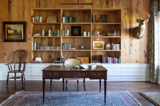 a farmhouse home office with a wood clad wall, built-in bookshelves in a niche, a vintage desk and some chairs plus a bold rug