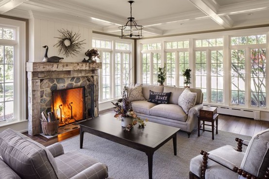 a neutral farmhouse sunroom with a stone clad fireplace, elegant furniture, black vintage tables and a vintage chandelier