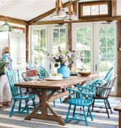 a bright farmhouse sunroom with stained wooden beams, a rustic wooden table, bright blue chairs and vintage pendant lamps