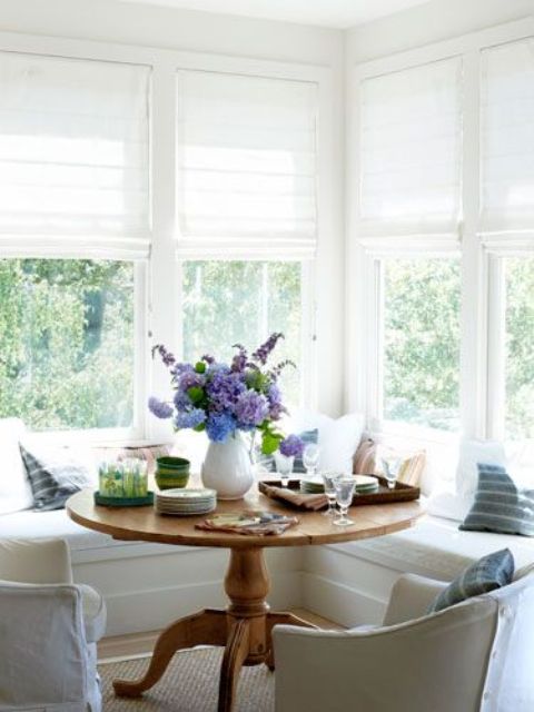 a neutral farmhouse sunroom corner with an L-shaped window seat and a rustic vintage table for having breakfasts here
