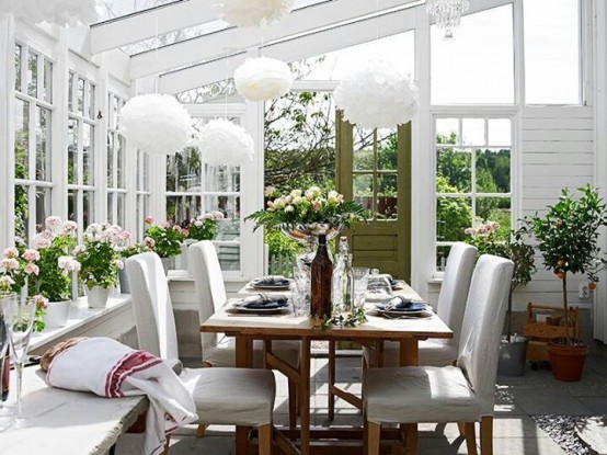 a white farmhouse sunroom with white furniture, a wooden table, fluffy pendant lamps and potted greenery and blooms is very cozy