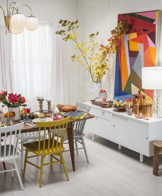 an eclectic dining room with a stained table, mismatching chairs, an oversized colorful geometric artwork, branches with fall leaves