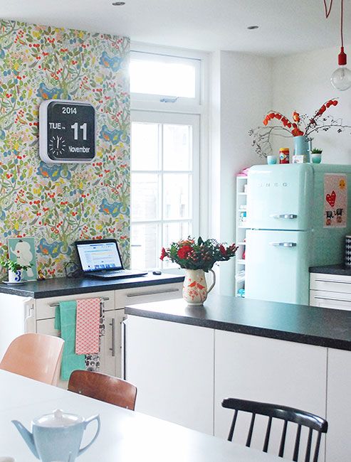 a catchy Scandinavian kitchen with floral wallpaper, a mint blue fridge, white cabinets, a small dining space with mismatching chairs