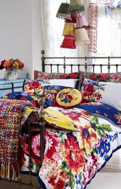 a bright boho bedroom with a light blue dresser, a forged bed with bold floral bedding, colorful pendant lamps and crochet blankets