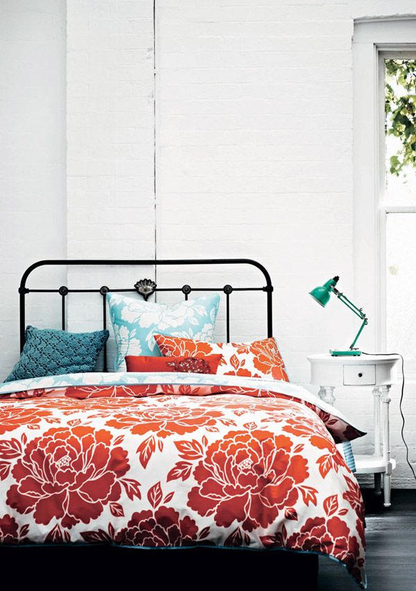 a white bedroom with a blakc forged bed and bold floral bedding, turquoise pillows, a white side table and a turquoise lamp