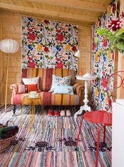 a bold sunroom with stained wooden walls and a ceiling, oversized floral accents, a bold striped sofa and a rug, a red stool and a side table