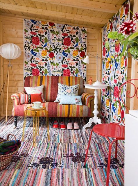 a bold sunroom with stained wooden walls and a ceiling, oversized floral accents, a bold striped sofa and a rug, a red stool and a side table