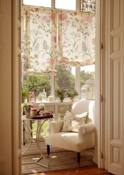 a small sunny space with floral shades, a long console table with decor, a white chair and a side table plus some potted plants