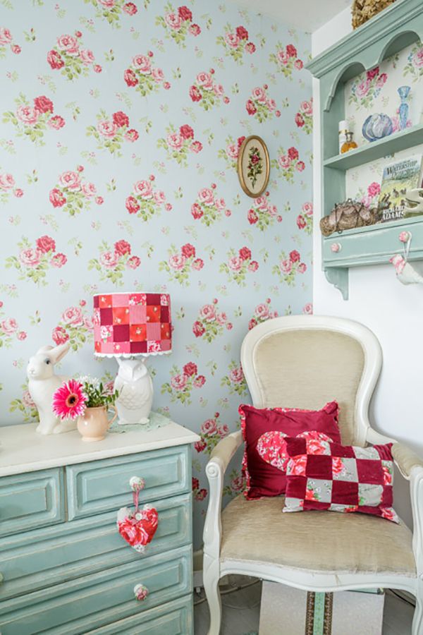 a cozy cottage space with floral wallpaper, a ligth blue shelving unit with decor, a matching dresser and a vintage chair