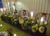 a gorgeous green Thanksgiving tablescape with a green tablecloth, green on the place settings, a box with pumpkins, apples, artichokes and candles is bold