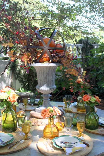a rustic Thanksgiving tablescape with green and amber glasses and bottles, green napkins and an urn with dried leaves and pumpkins