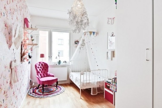 a whimsical and exquisite kid's room with a blush floral accent wall, shelves, a white bed with bedding, a pink chair and a whimsical floral chandelier
