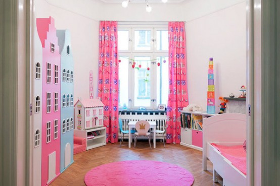 a colorful kid room with large dollhouses, a bed with pink bedding, a hot pink rug, a working space by the window with pink curtains