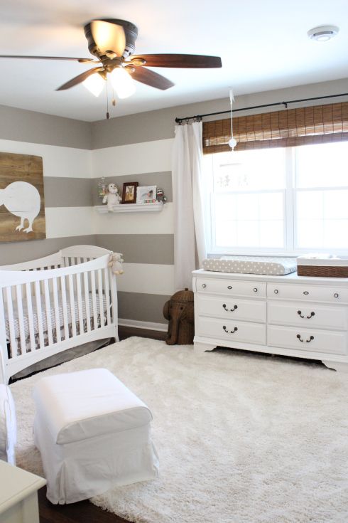 a neutral farmhouse nursery with a striped accent wall, white vintage furniture, a white faux fur rug, woven shades, neutral bedding