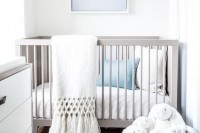 a white gender-neutral nursery with white and grey furniture, white and blue bedding, toys and an artwork is serene and airy