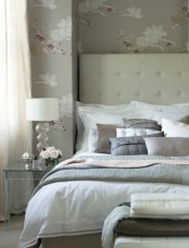 a grey glam bedroom with a white bed, printed pillows, floral wallpaper, a glass nightstand and lots of blankets