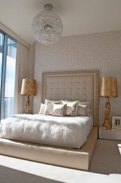a refined neutral bedroom with catchy wallpaper, a large creamy upholstered bed, neutral and printed bedding, gold table lamps, a floral chandelier is cool