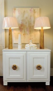 a small yet chic cabinet with beautiful gold knobs, with table lamps with gilded bases and pink lampshades plus a gold and pink artwork