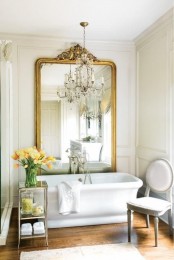 a beautiful vintage-inspired bathroom with an oversized mirror in a gilded frame, a white chair, a tub and a mini glass side table with blooms
