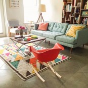 a bright mid-century modern living room with an aqua sofa, bright pillows, a colorful printed rug, a coffee table and rocker chairs, a large bookcase and floor lamps