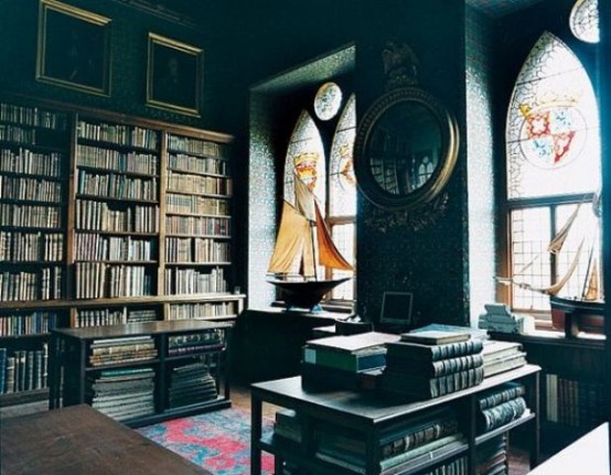 a Gothic library with mosaic windows, bookshelves covering the whole wall, a gallery wall, a couple of tables with books and a desk plus ships on the windowsills