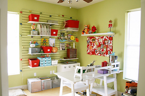 a green home office with open shelves, bright red decor, a white desk with colorful accessories and lots of natural light