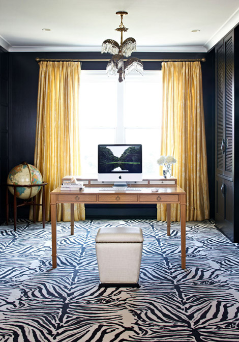 a chic home office with black walls and a built-in storage unit, a light-stained desk and a white leather stool, a zebra-print rug plus a large vintage globe