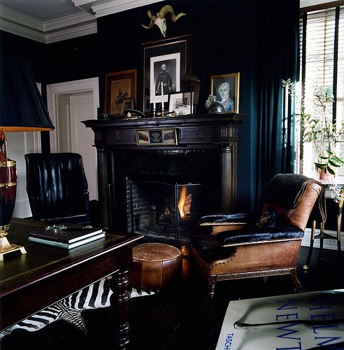 a refined moody home office with black walls, a large fireplace with a black mantel, a refined black desk and a black and a brown leather chair plus greenery