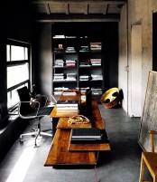 a moody industrial home office with black and neutral walls, a large storage unit, a stained desk and a black leather chair, some artwork and a window with natural light