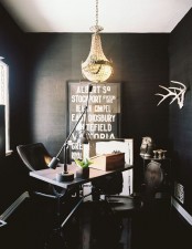 a sophisticated black home office with a small and laconic desk, a black leather chair, a gorgeous crystal chandelier and a statement artwork plus antlers and some more elegant decor