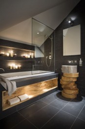 a dark home space with a tub and niches with candles over it, a unique vanity of tree slices, a wooden niche for towels and built-in lights welcomes in