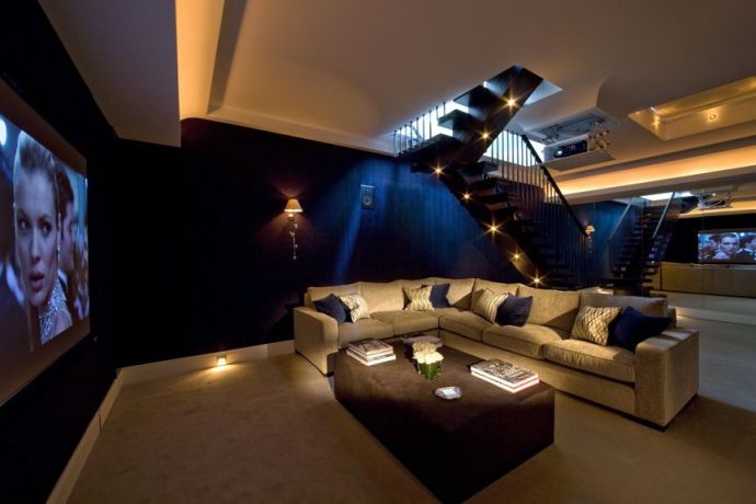 a pretty living room turned into a home theater, with a large screen, a corner sofa, a brown pouf and built in lights around