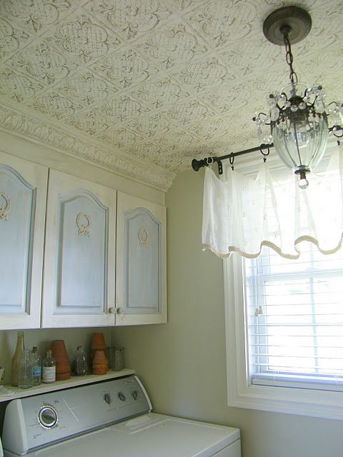 a neutral vintage inspired space with a white tin tile ceiling, neutral cabinets, a crystal chandelier and some curtains