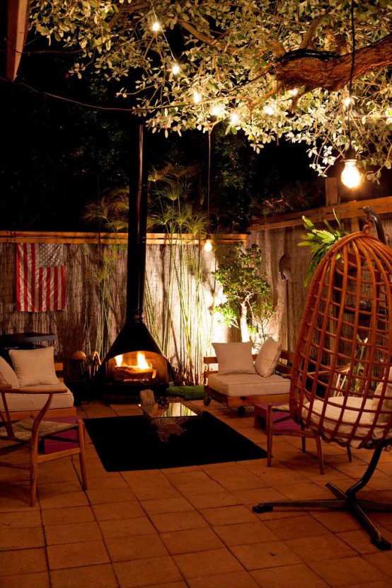 a cozy outdoor living room with a black Malm fireplace, neutral seating furniture, a rug, an egg pendant chair and pendant bulbs