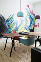 a contemporary dining room with a trestle dining table and colorful chairs and colorful pendant lamps that echo with the accent graffiti wall