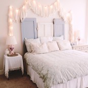 a vintage meets shabby chic bedroom in neutrals, with a bed and pastel bedding, a headboard formed of old doors and tassels accented with lights that add coziness to the space and make it more lit up
