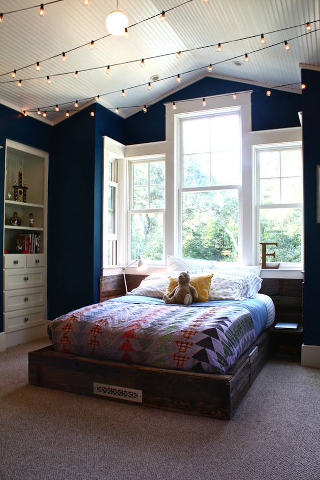 a farmhouse bedroom with navy walls, wihte built in furniture, a stained bed with colorful bedding and string lights over the whole room