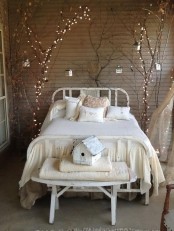 a shabby chic bedroom with a planked taupe accent wall, a white bed and bench, branches and trees with string lights for a bright accent