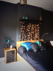 a moody black bedroom with light-stained furniture, black and blue bedding, a shelf that holds string lights that form a wardrobe