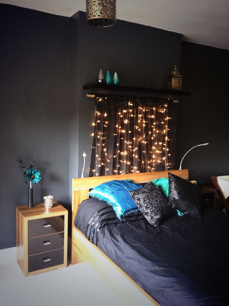 a moody black bedroom with light stained furniture, black and blue bedding, a shelf that holds string lights that form a wardrobe