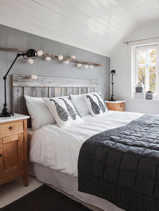 a modern rustic bedroom with a grey accent wall, a piece of driftwood with string lights covering it, a bed with black and white bedding, stained nightstands and black lamps
