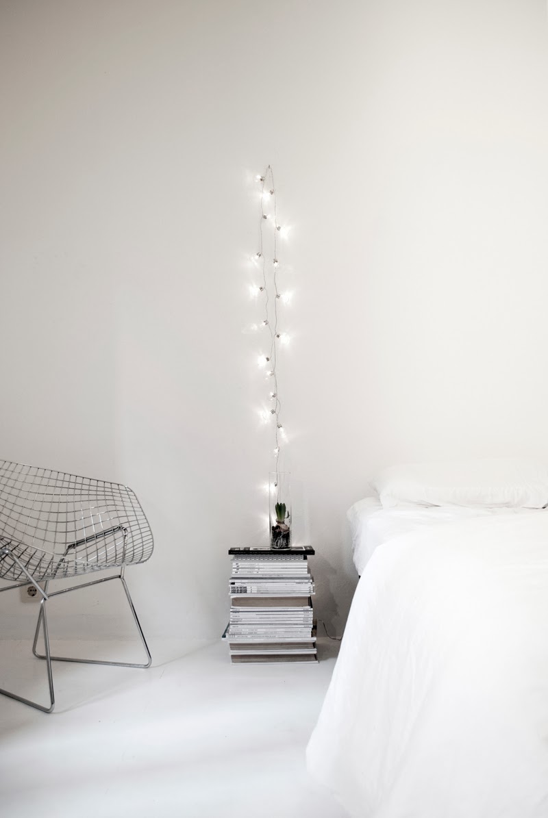 a minimalist Scandinavian bedroom with a low bed with neutral bedding, a metal chair, a stack of magazines and string lights hung over them as a light source, an alternative to a usual table lamp