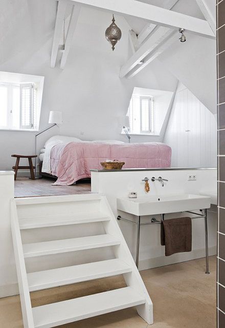 a white attic loft bedroom with wooden beams and Moroccan pendant lamps, a bed with white and pink bedding, a nightstand and a bathing space downstairs