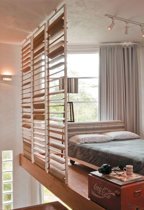 a loft bedroom featuring a large window, a neutral curtain, a comfy bed and a chest as a nightstand separated from the rest of the house with blinds